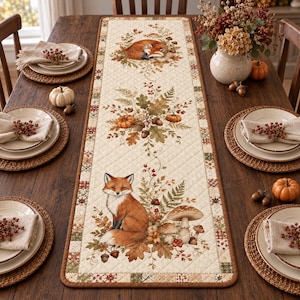 May include: A quilted table runner featuring a cream-colored background with a repeating autumn-themed print. The design includes illustrations of a sleeping fox, a sitting fox, pumpkins, acorns, mushrooms, and foliage in shades of orange and brown.