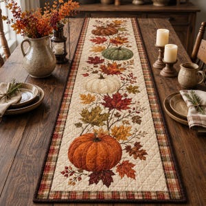 Puede incluir: Un camino de mesa acolchado rectangular con un estampado central de calabazas en naranja, blanco y verde, rodeado de hojas de otoño en tonos rojos, dorados y marrones. El camino tiene un centro color crema y un borde a cuadros en tonos tierra.