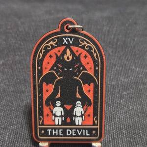 May include: A wooden tarot card keychain featuring "The Devil" card. The design includes a black silhouette of a devil with wings, two small figures, and the text "THE DEVIL". The card is framed in red and gold.