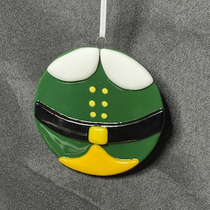 May include: A round Christmas ornament featuring a green elf costume design. The ornament has white collar details, a black belt, and a yellow buckle and collar. The ornament is suspended by a white ribbon.