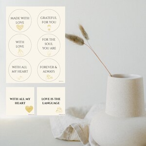 Könnte beinhalten: Eine Sammlung cremefarbener Aufkleber mit goldenem Text und Illustrationen. Die Aufkleber zeigen Sätze wie "Made with Love", "Grateful for You" und "Love is the Language". Im Hintergrund befindet sich eine minimalistische Vase mit getrockneten Blumen.