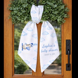 Puede incluir: Decoración de puerta de baby shower de tela blanca con nubes azules y una ilustración de avión. El texto dice "Sophia's baby shower" y "August 12, 2025". La decoración está colgada en una puerta de madera.