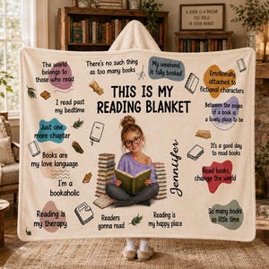 May include: A cream-colored hooded blanket featuring the text "THIS IS MY READING BLANKET" and "Jennifer," with a central image of a child reading a book. The design includes various book-themed quotes, illustrations of books, and colorful abstract shapes.