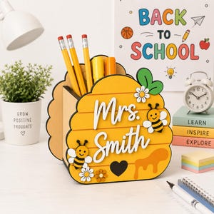 May include: A yellow wooden desk organizer shaped like a beehive, featuring the text "Mrs. Smith" in white script, two cartoon bees, white flowers, and a black heart. The organizer holds three yellow pencils and two markers.