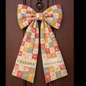 May include: A patchwork fabric bow, with floral squares in red, yellow, blue, and green, hanging on a wooden door. One tail reads "GRANDMA", the other "Emma • Olivia • Noah". The bow is a decorative item, suitable for a door or wall.