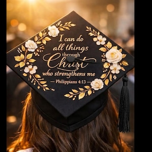 Op de afbeelding: Zwarte afstudeerhoed versierd met gouden bladeren, witte bloemen en de tekst "I can do all things through Christ who strengthens me - Philippians 4:13". Een zwarte kwast hangt aan de hoed.