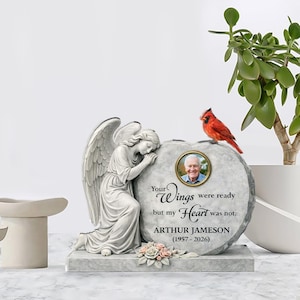 Può includere: Una lapide commemorativa con la foto di un uomo, una scultura di angelo e un cardinale rosso. La pietra è incisa con la frase "Your Wings were ready but my Heart was not." e il nome "ARTHUR JAMESON (1957-2026)."