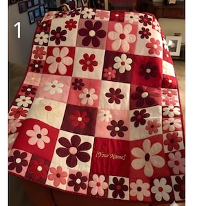 May include: A handmade quilt featuring a floral pattern in shades of red, pink, and white. The quilt is composed of square blocks, each adorned with appliqued flower designs. The text "[Your Name]" is visible on the quilt.