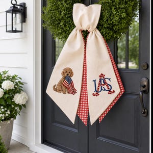 May include: Cream-colored door swag with a red and white gingham trim. Features an embroidered brown dog holding an American flag and the letters "US" in blue script. A decorative accent for a front door.