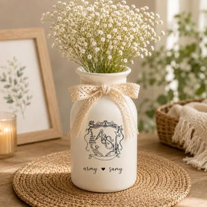 Könnte beinhalten: Eine weiße Keramikvase mit cremefarbener Spitzenschleife, gefüllt mit weißen Blumen. Die Vase hat ein schwarzes Wappen-Design mit den Initialen "A" und "S" und den Worten "army" und "samy". Die Vase steht auf einer gewebten Matte.