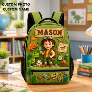 Könnte beinhalten: Ein grüner Rucksack mit einer 3D-Figur eines Entdeckers, der eine Lupe hält, umgeben von Dinosaurierfiguren, einem Vulkan, einem Kompass und Fossilien. Der Name MASON ist auf einem rechteckigen Aufnäher oben auf dem Rucksack aufgedruckt.