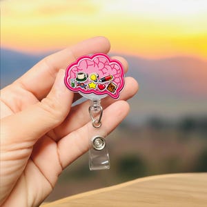 Puede incluir: Un portatarjetas rosa en forma de cerebro con varios iconos coloridos, incluyendo una taza de café, una bombilla y un bolígrafo. El portatarjetas tiene una correa de plástico transparente y un clip de metal.