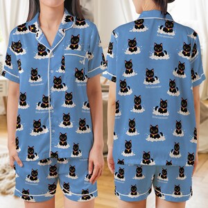 Könnte beinhalten: Hellblaues Pyjama-Set mit einem sich wiederholenden Muster aus schwarzen Katzen auf weißen Wolken. Das kurzärmelige Oberteil und die Shorts haben weiße Paspeln und den Text "Peter Alexander". Die Katzen tragen goldene Halsbänder.