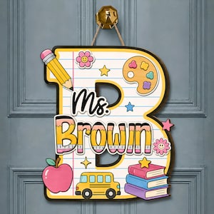 May include: A decorative wooden wall sign shaped like the letter B with a lined notebook paper pattern. It features the text Ms. Brown, a yellow pencil, a paint palette, a pink apple, a yellow school bus, and a stack of three colorful books.