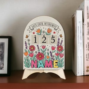 May include: A wooden retirement countdown calendar displaying "DAYS UNTIL RETIREMENT" above the numbers 125. The calendar is decorated with flowers and the word "NAME". It stands on a wooden easel, with a floral design.