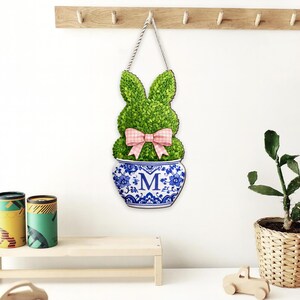 May include: A wooden Easter bunny wall decoration. The green bunny has a pink gingham bow and sits in a blue and white floral pot with the letter "M". It hangs from a rope and is mounted on a wooden rack.
