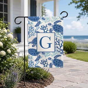 May include: A garden flag featuring a blue and white sea turtle and coral pattern. The flag has a white square with the letter "G" in the center. The flag is displayed on a black metal stand in a garden setting.