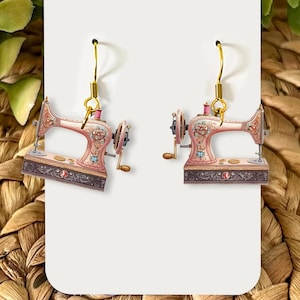 May include: A pair of earrings featuring a vintage sewing machine design. The earrings have a pink and brown color scheme with gold-colored hooks. The sewing machines are detailed with floral patterns and decorative elements, evoking a retro aesthetic.