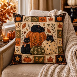 May include: A square throw pillow featuring a patchwork-style border of pumpkins, leaves, and stars. The center panel displays three black cats interacting with pumpkins, including one cat wearing a plaid bow, set against a cream background.