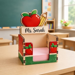Könnte beinhalten: Ein Schreibtisch-Organizer aus Holz mit einem roten Apfel und einem gelben Bleistift als Ausschnitt oben, mit dem Text Ms. Sarah auf einer horizontalen Leiste. Die Seiten sind mit roten und grünen Apfelgrafiken verziert und halten rosa Papier.