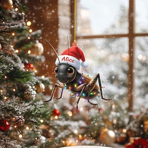 May include: A Christmas ornament featuring a cartoon cricket wearing a Santa hat with the name "Alice" and a string of colorful lights. The cricket is black and brown, with large eyes. The ornament hangs in front of a Christmas tree.