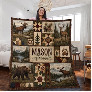 Puede incluir: Una colcha rectangular con diseño de patchwork que presenta imágenes de un alce, un ciervo, un oso, pinos, montañas y una huella de oso. El panel central muestra el nombre Mason Alexander en letras de color marrón oscuro sobre fondo claro.