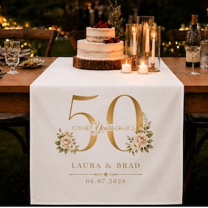 May include: A white fabric table runner featuring a large gold number 50, the word Years, and floral illustrations. The text LAURA & BRAD and the date 06.07.2028 are printed in gold below the number. The runner is displayed on a wooden table.