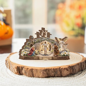 Può includere: Statuina religiosa raffigurante la risurrezione di Gesù, con una figura luminosa che emerge da una tomba. La scena include croci, angeli e figure in abiti, su una base di legno. Il testo "He is Risen" è visibile.