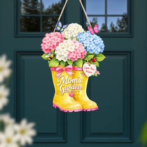 Może przedstawiać: Ozdobny szyld na drzwi z parą żółtych kaloszy wypełnionych różowymi, białymi i niebieskimi kwiatami hortensji. Buty mają różową kokardę i napis "Mom's Garden". Etykieta w kształcie serca mówi "Love You Mom". Obecne są również motyle i pszczoła.