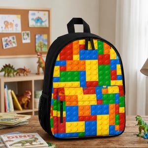 May include: A black backpack featuring a vibrant, multi-colored pattern of interlocking plastic building blocks in red, blue, yellow, and green. The bag includes a main zippered compartment and a smaller front pocket, both with black zipper pulls.