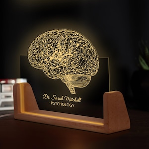 May include: A lighted brain-shaped sculpture with the name "Dr. Sarah Mitchell - PSYCHOLOGY" on a black background. The brain design is illuminated in a warm yellow color, set in a wooden base with a glowing light.