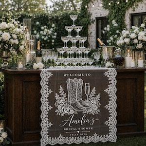 May include: A dark brown table runner with white lace trim featuring a printed design of two cowboy boots, a champagne flute, and roses. The text reads: Welcome to Amelia's Bridal Shower 10.11.2025. It is displayed on a wooden table.