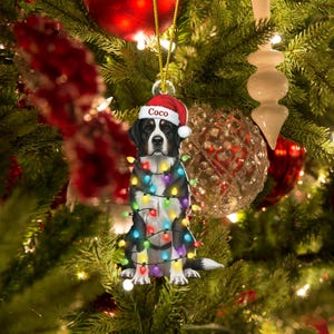 May include: A Christmas tree ornament featuring a dog wearing a Santa hat with the name "Coco" and wrapped in colorful Christmas lights. The dog is black, white, and brown, and the ornament hangs from a gold string.