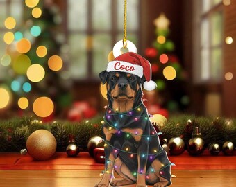 May include: A Rottweiler dog decoration wearing a Santa hat with the name "Coco" and wrapped in colourful Christmas lights. The ornament hangs in front of a blurred Christmas tree and festive decorations.