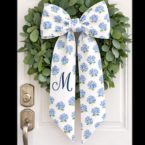 Peut inclure: Un nœud blanc avec un motif floral d'hortensias bleus et la lettre "M" bleu marine est attaché à une couronne verte. Le nœud est suspendu à une porte blanche avec une poignée dorée.