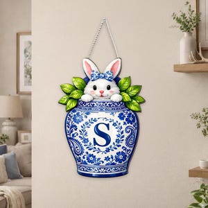 Könnte beinhalten: Blau-weiße Wanddekoration mit einem Hasen, der aus einer Vase schaut. Die Vase hat ein Blumenmuster und den Buchstaben "S". Der Hase trägt eine blaue Schleife und ist von grünen Blättern umgeben. Aufgehängt an einer Schnur.