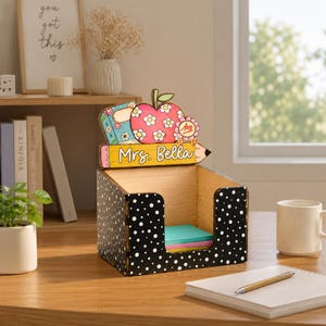 Könnte beinhalten: Ein schwarzer Schreibtisch-Organizer aus Holz mit weißen Punkten und einem dekorativen Aufsatz, der einen Blumenapfel, ein Buch und einen gelben Stift mit der Aufschrift "Mrs. Bella" zeigt. Die offene Box enthält bunte Haftnotizen.