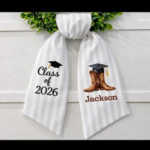 May include: White and gray striped graduation sash with the text "Class of 2026" and a graduation cap graphic. The other side features brown cowboy boots with a graduation cap and the name "Jackson".