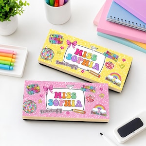May include: Two rectangular pencil cases, one yellow and one pink, featuring the name MISS SOPHIA in colorful bubble letters. The cases are decorated with stickers of a disco ball, rainbow, apple, pencil, bow, and text reading LET'S LEARN TODAY, YOU'RE ENOUGH, YOU'RE AMAZING, and YOU'RE LOVED.