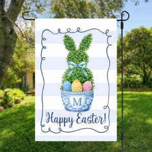 May include: A garden flag with a blue and white striped design. The flag displays a green topiary rabbit with a blue bow, a blue and white bowl with three Easter eggs, and the words "Happy Easter!"