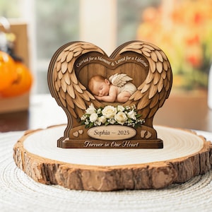 Könnte beinhalten: Ein hölzernes Gedenkstück mit einem schlafenden Baby mit Engelsflügeln, eingebettet in einen herzförmigen Rahmen aus geschnitzten Flügeln. Der Text lautet "Carried for a Moment & Loved for a Lifetime", "Sophia - 2025" und "Forever in Our Heart."