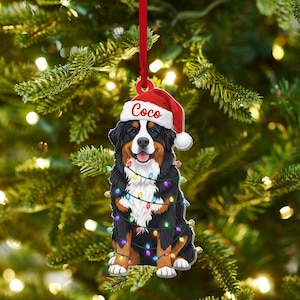 May include: A Christmas ornament featuring a Bernese Mountain Dog wearing a Santa hat with the name "Coco" and string lights. The dog is black, brown, and white, with a red ribbon for hanging. The background is a blurred Christmas tree.