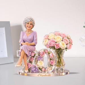 May include: A figurine of a woman in a lavender dress, seated next to a floral arrangement and a '70' display. The '70' is adorned with roses and other flowers. A vase of pink and yellow roses sits nearby. The base is inscribed with the name "Elizabeth".