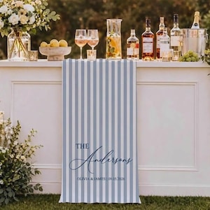 May include: A white bar with a blue and white striped fabric panel. The panel reads "The Andersons" with the names Olivia & James and the date 09.05.2026. The bar is set with drinks, glasses, and flowers.