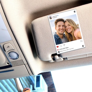 May include: A car visor with a photo of a smiling couple, resembling an Instagram post. The post has the usernames @emma and @jason, with a heart icon and the text "1,000,000 likes." The caption reads "Drive safe, my love."