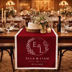 Puede incluir: Camino de mesa burdeos con borde blanco, con un diseño de corona floral con las iniciales "E & L" y los nombres "Ella & Liam". También se muestra la fecha "09.14.2025". La mesa está preparada para una boda.