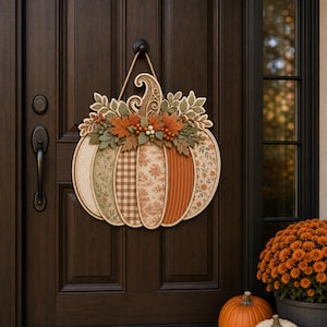 May include: A decorative wooden pumpkin door hanger featuring five vertical segments with varied patterns including gingham, floral, and solid orange stripes. The top is adorned with layered wooden leaves, berries, and a curled stem, all suspended by a tan rope.