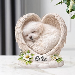 Könnte beinhalten: Cremefarbene Skulptur eines schlafenden weißen Hundes, der in herzförmigen Engelsflügeln liegt. Der Sockel trägt den Namen "Bella" und Blumenschmuck. Ein Andenken.