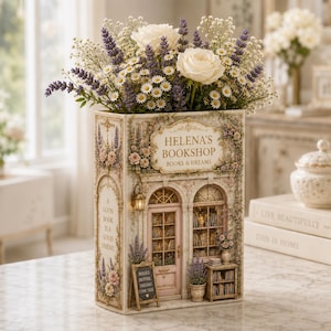 May include: A decorative book-shaped vase featuring a detailed illustration of a storefront labeled Helena's Bookshop Books & Dreams, filled with white roses, purple lavender, and small white daisies, with text A Good Book Is A Good Friend.