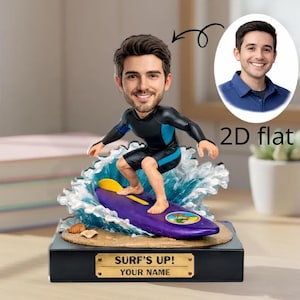 May include: A personalised surfer figurine, featuring a smiling face, riding a purple surfboard on a wave. The figurine wears a black and blue wetsuit. The black base has a gold plaque reading "SURF'S UP! YOUR NAME".